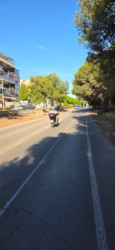 A slightly rotated, medium-angle shot captures a motorcycle moving on a road in Canet d'en Berenguer, Spain. The motorcycle is white with a single rider. The road, marked by a white line, extends into the distance with shadows cast onto its surface. To the left of the road, a multi-story residential building is visible, with balconies and solar panels. Trees line the roadside. The sky is a clear blue. Other parked cars can be seen along the roadside. The location is identifiable as Canet d'en Berenguer due to the typical architecture and landscape of the area.