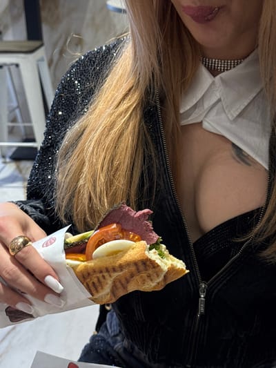 An individual is depicted holding a large sandwich, presumably in a casual dining establishment in Asnières-sur-Seine, France. The person's torso and lower face are visible, showing light brown hair. They wear a black zippered jacket adorned with reflective embellishments, a white collared shirt, and a dark undergarment revealing cleavage. A multi-row choker necklace is present, and a dark tattoo is visible on the upper right chest. The person's manicured left hand, wearing a gold-toned ring, holds a partially wrapped baguette-style sandwich. The sandwich contains slices of reddish meat, tomato, hard-boiled egg, and green leafy vegetables. The individual's lips, coated in dark pink lipstick, are slightly parted with a hint of the tongue visible. The background is blurred, showing a light-colored surface and a metallic chair component.