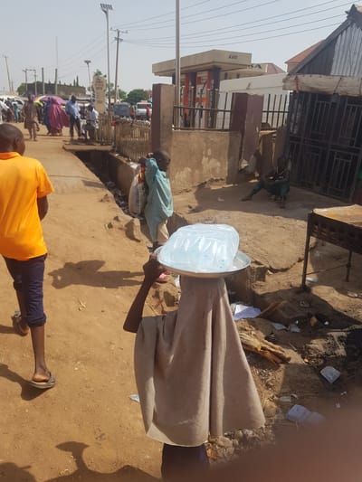 This vertical image depicts a bustling street scene in Tukur Tukur, Nigeria, under bright daylight.
In the immediate foreground, a person, mostly covered by a long, light brown garment, carries a large metal tray filled with numerous clear plastic bags of water on their head. Their right arm is visible, holding a stick. To the left of this central figure, another person in a bright orange t-shirt and dark trousers walks away from the camera. A young boy in a light blue shirt stands nearby, facing the water carrier.
The ground is unpaved, consisting of dry, light brown earth, uneven in texture, with some scattered debris and waste visible. In the background, a busy street stretches into the distance, lined with various buildings and structures, some with corrugated iron roofs. Several people are visible walking or standing along the street. Text visible on a building includes "AGO" and part of "KA". Power lines traverse the scene, and a solar-powered streetlight can be seen. Vehicles, including a truck, are present further down the road. The strong, direct sunlight creates sharp shadows, indicating a sunny midday or early afternoon.