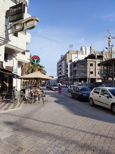 This media file depicts a street scene in Tunisia, likely during daytime given the clear blue sky and bright sunlight casting shadows. The image captures an outdoor cafe with tables and chairs set up on a paved sidewalk. A large beige umbrella shades some of the tables.
In the foreground, several cars are parked along the street, including a white Fiat in the right foreground. Further down the street, more vehicles are visible, along with some palm trees. On the left side of the street, a building features signage for "RODENSTOCK" and "Optylab," suggesting an optician's shop, and a large novelty pair of glasses as a sign. Another sign for a "PHARMACIE" with a neon green outline is also visible.
In the background, construction is underway on a building, with a crane prominently visible. Other buildings of various architectural styles line the street. A few people are visible walking along the sidewalk and street, including a woman in a pink dress. The overall atmosphere appears calm and ordinary, suggesting a typical day in a Tunisian town or city. There are no overt signs of distress or celebration.