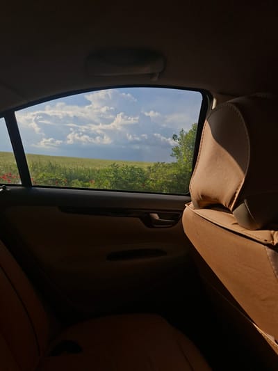 The image depicts a view from inside a car. The interior is beige and shows car seats and trim. Through the car window, there is a view of a field with green grass and red flowers, with a cloudy blue sky above. The location is identified as Pobeda, Bulgaria. No people are visible.