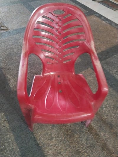 A red plastic chair is placed on a patterned outdoor floor. The chair has a slatted backrest and armrests. The floor appears to be made of concrete tiles with a dark grey and light grey pattern, with some areas of dirt and debris. The lighting suggests it is daytime, with shadows cast on the floor. The image is taken from a high angle, looking down at the chair. The chair is the primary subject of the image, and it is positioned in the center. The background shows a continuation of the patterned floor and a blurry area that may be a wall or another part of the outdoor environment. The scene appears to be in an outdoor public space, potentially a walkway or patio area. The location is Pekanbaru, Indonesia.