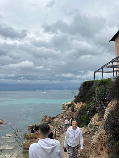 The image depicts a coastal scene in Llucmajor, Spain, on a cloudy day. Turquoise water meets the rocky cliffs, and a few sailboats dot the horizon. A metal structure sits atop a building near the cliff's edge. Several people are visible on a path along the cliff. One woman in a white top and patterned pants is facing the camera, while another person in a white hoodie is partially visible in the foreground. A wooden railing runs along part of the path.