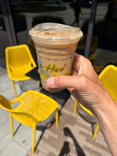 A light-skinned human hand, exhibiting visible veins and a minor blemish on the thumb's knuckle, firmly holds a clear plastic cup filled with an iced beverage, consistent with an iced latte or coffee. The cup features a yellow "THE Hive" logo, accompanied by the words "HEALTHY," "HAPPY," "KIND," and "TREATS," along with a bee icon and blue and yellow floral illustrations. The beverage is capped with a clear plastic dome lid, showing condensation and liquid residue. The scene is outdoors under direct sunlight, evidenced by strong shadows cast on the ground. In the background and mid-ground, multiple bright yellow, perforated metal chairs are arranged on a patio paved with light-toned rectangular tiles and some reddish-brown brick-patterned sections. A dark, reflective glass surface, likely a storefront window, occupies the upper background, showing blurred reflections of distant figures and structures. The location is identified as "The Hive: Healthy Food & Coffee," situated at 4333 Admiralty Way, Marina del Rey, United States.