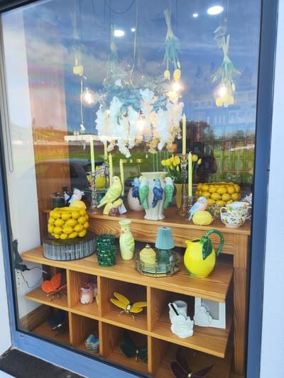 A window display in Ponta Delgada, Portugal, showcases an array of decorative objects and furnishings. The items are arranged on a multi-tiered wooden shelf unit. On the upper shelf, there are decorative birds, candle holders, vases (one with bird motifs), faux lemons, and teacups. The lower shelves feature ornamental butterflies, candles, and other decorative items. Above the shelves, floral arrangements and hanging light fixtures are suspended. Reflections in the glass show the cityscape and sky, indicating the location is likely a shop or gallery in an urban area.