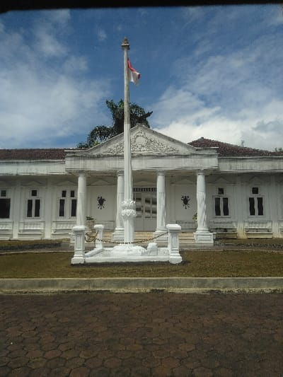 The image displays a white, colonial-style building in Palasari, Indonesia, under a blue sky with scattered white clouds. A tall, white flagpole stands centrally in front of the building, from which the red and white flag of Indonesia flies. The building features a prominent central portico supported by four white classical columns, topped with an ornate pediment. White-framed windows are visible on either side of the entrance, and two dark, star-shaped wall decorations are mounted on the facade. The flagpole's base is white and ornate, surrounded by white bollards linked by black chains. The immediate ground around the flagpole is sparse green grass, bordered by a grey concrete curb. The foreground consists of a dark red-brown paved surface composed of hexagonal bricks. A large green tree is partially visible behind the building on the left. No individuals, specific actions, or interactions are present in the scene.