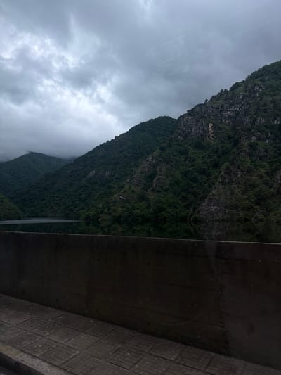 The image shows a view of a mountain landscape partially obscured by low-hanging clouds, possibly taken from inside a vehicle or structure with a window. The mountain is covered in dense greenery, likely vegetation or forest. A section of a concrete structure with a visible pattern is in the foreground, partially obstructing the view. The sky is overcast with grey clouds, suggesting a cloudy or rainy day. The location is not visibly discernible as Krichim, Bulgaria, based on the content of the image.