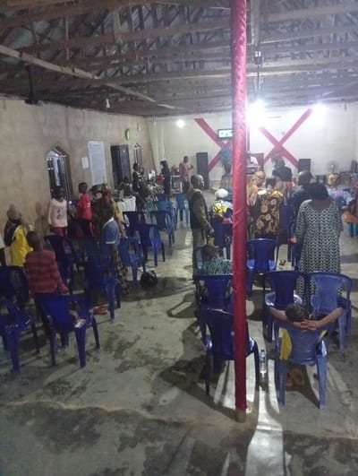 A large group of people gathers inside a dimly lit building, likely a church, with a wooden ceiling. Several rows of blue plastic chairs fill the space, some occupied, others empty. The scene is centered around a raised platform adorned with red crosses. A red-covered post stands prominently in the foreground, casting a shadow. The building appears modest in design, with simple windows. Some individuals are standing while others are seated.