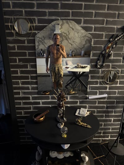 The image captures a man taking a selfie in a room with dark brick walls. The man, shirtless and wearing patterned shorts, is standing in front of a large mirror that reflects the room. The mirror also shows a glimpse of a kitchen area with cabinets and appliances. On a round black table in the foreground, there are various objects, including a Tibetan singing bowl, a decorative figurine, some stones, a small glass globe, and a deck of cards. The room has a modern aesthetic with a black tiled floor. The overall impression is of a personal space, possibly a home or studio, in the city of Vol'noe, Russia.