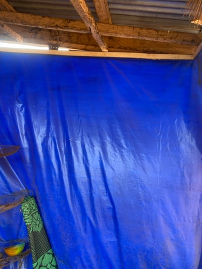 The image captures an interior view of a provisional structure located in the city of Okura, Nigeria. The primary visual element is a large, crinkled blue plastic tarpaulin, which forms the main wall, extending from the upper frame to the bottom and across most of the width. Above the blue tarp, a segment of the roof structure is visible, comprising multiple rough-hewn wooden beams or rafters supporting a corrugated metal sheet roof. A gap between the top edge of the tarp and a horizontal wooden beam allows some ambient light to penetrate. In the upper right corner, a small amount of dried organic material, possibly thatch or leaves, is partially visible.
In the lower left quadrant, several objects are present against the blue tarp. Multiple light brown, coarse, net-like or woven items are partially visible, suggesting storage or hanging implements. Below these, a piece of green fabric or a mat, patterned with black floral or geometric designs, leans vertically against the blue tarp. Adjacent to the bottom-most woven item and the green mat, a small, multi-colored bowl, exhibiting a gradient from yellow to orange, is resting. No individuals or active interactions are depicted in the scene.