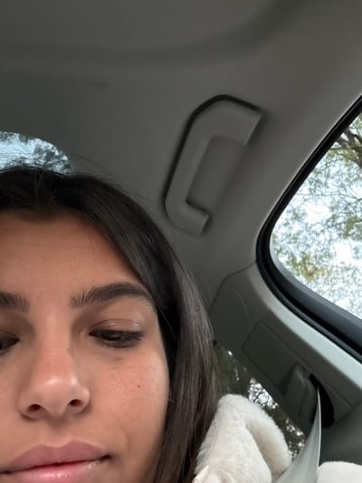 An individual is seated within the passenger compartment of a vehicle in Lawrence Township, United States. The lower half of a female's face, dark brown hair, and upper torso are visible, positioned towards the left and center of the frame. She has a medium-dark skin tone and her eyes are cast downwards and slightly to the right. A white, possibly faux fur, garment covers her right shoulder and chest area, and a silver-colored seatbelt buckle and strap are visible on her lower right. The vehicle's interior features a light gray headliner and an overhead grab handle. Through the vehicle's side window, tree branches are visible against a light sky, indicating an outdoor, daylight environment.