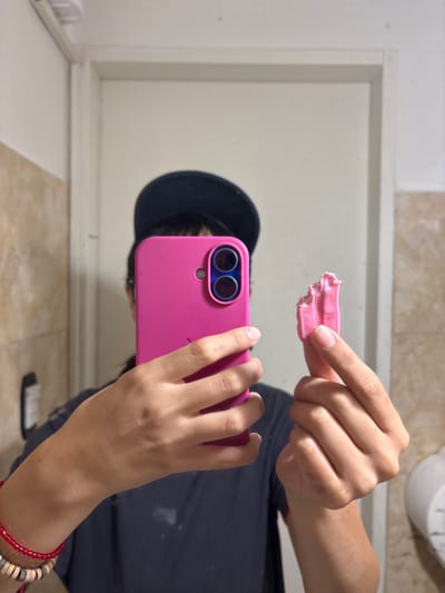 A person holds a pink smartphone in front of a mirror. In the mirror's reflection, the person is wearing a black hat. The person uses their other hand to hold a piece of pink chewing gum in the air between their fingers. The scene appears to be a bathroom. The smartphone is a bright pink color with dual cameras. The bracelets of red and brown beads adorn the person's wrist.