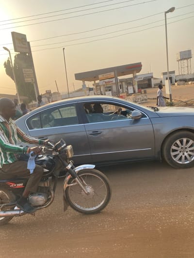 The image captures an outdoor scene at a TotalEnergies gas station in Nigeria during either dawn or dusk, characterized by a warm, golden sky. In the foreground, a person is seated on a black Yamaha AX100 motorcycle, wearing a striped green, white, and brown shirt, dark pants, and a light blue face mask pulled down to their chin.
Adjacent to the motorcycle and slightly behind it, a grey sedan car is parked. A person is visible inside the car, and another individual, dressed in a light-colored long-sleeved shirt, stands by the front passenger side, possibly interacting with the vehicle or gas station staff.
The background prominently features the TotalEnergies gas station, displaying various brand names such as "Bonjour," "QUARTZ," and "Valvoline." A price board clearly shows fuel prices in Nigerian Naira: "PMS N555," "AGO N580," and "DPK N580." Utility lines are visible against the soft orange sky, and in the far distance, a water tower or similar structure can be seen. The ground beneath the vehicles appears to be a dusty road surface. The scene depicts a calm, everyday moment, likely involving waiting at the gas station.