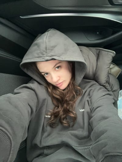 The image displays a single individual, identified as a young adult female, positioned inside a vehicle interior. The location is specified as Stara Pazova Municipality, Serbia. The female wears a dark grey hooded sweatshirt with the hood up, obscuring the upper portion of her head. Visible on the front of the sweatshirt are embroidered white texts: "Do Shit" on the left chest and a partial "You Bel" on the right. Long, wavy brown hair extends from beneath the hood and flows over her shoulders. She has light-colored eyes and appears to be looking directly into the camera, consistent with a self-portrait capture. Behind her shoulders, a dark grey puffer jacket is draped, resting against the vehicle's seating or interior paneling. The immediate background reveals the vehicle's dark-toned interior, including a textured panel to the left of the subject and an overhead component (headliner). A light-colored object is partially visible behind the puffer jacket, positioned to the right of the subject's head.