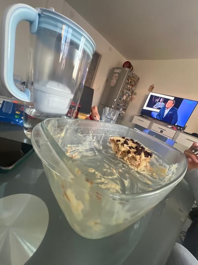 The image depicts a kitchen scene. A glass baking dish sits on a gray countertop with a partial piece of cake/dessert inside. A water filter pitcher with a blue handle is also present on the counter. In the background, there is a refrigerator covered with magnets and a television showing a person on screen. The location is presumed to be in Benningen, Germany, based on available information.