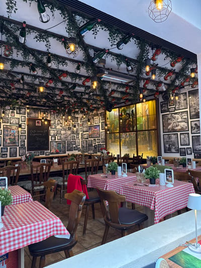 The image captures the interior of a restaurant in Cochem, Germany. Tables are covered with red and white checkered tablecloths, set with plants and menus. Chairs are wood and dark colored. The walls are adorned with rows of framed black and white photographs and vintage posters. The ceiling on the left is decorated with wine bottles, greenery, and wire-frame light fixtures. A chalkboard on the wall lists menu items, including "Cafe Germa," "Cappuccino," and "Jupiler." There is a large stained glass window visible in the background. The overall setting is warm and inviting with vintage decor.