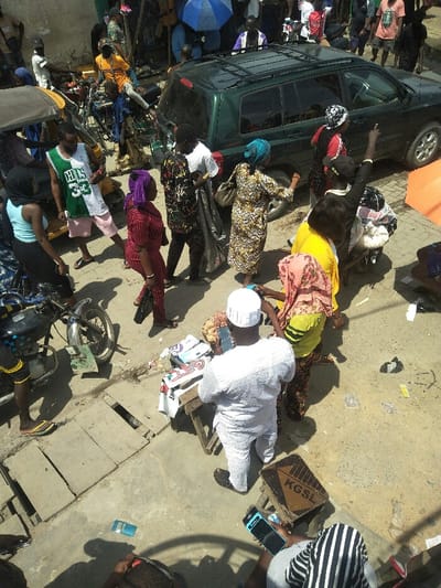 The image provides an elevated view of a bustling street scene in Ajah, Nigeria, under direct sunlight, indicated by prominent shadows. The ground consists of uneven concrete slabs, visible manhole covers, and scattered litter including plastic bottles and papers.
Numerous individuals, appearing to be adults of varying ages, are present and actively moving or congregating. A dark green SUV, resembling a Toyota Highlander, is centrally positioned, either parked or slowly navigating through the crowd. To the left, a black motorcycle is stationary.
Key individuals and objects include:
* In the foreground, a male, dressed in a white traditional tunic (agbada-like) and a white cap, stands with his back to the viewer, holding a mobile phone. He faces a small, rudimentary wooden vendor's table displaying various packaged goods and textiles.
* Adjacent to him, a female in a yellow top, patterned skirt, and a pink headscarf is also holding a mobile phone.
* Near the SUV, a female in a yellow and brown patterned dress and a blue headscarf is observed. Beside her, a male in a white t-shirt and dark patterned trousers carries a large dark bag.
* On the left, a male wearing a green and white Boston Celtics jersey with the number "33" and light pink shorts stands near the motorcycle.
* In the lower right, an individual is partially visible, seated or crouching, holding a mobile phone next to a wooden crate marked "KGSL".
* In the background, further pedestrians are visible, some standing under a prominent blue umbrella, and a portion of an orange umbrella is seen on the right side of the frame.
The overall environment suggests an active urban market or thoroughfare with frequent pedestrian and vehicular traffic.