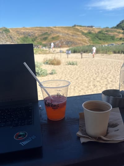 A laptop sits on a dark surface with a colorful drink in a plastic cup and a white paper cup of coffee beside it. A straw pokes out of the colorful drink, which has a "Veleka beach" logo. The background shows a sandy beach with a few people and a grassy, hilly landscape under a clear blue sky.