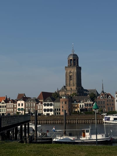 The image shows a waterfront view of Deventer, Netherlands. In the background, a prominent clock tower, part of the Grote of Lebuïnuskerk, stands tall against a clear blue sky. The tower features gothic architectural elements and a distinctive clock face. To the left and right of the tower, a row of historic buildings with red-tiled roofs lines the waterfront. In the foreground, a wooden pier or walkway extends into the water, with several boats docked nearby. One white boat in the center has a Dutch flag flying from its stern. The water is calm, and the scene suggests a sunny day. The grassy bank in the immediate foreground indicates the viewpoint is from land.