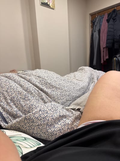 In a bedroom, the camera is positioned at a low angle, focusing on a floral-patterned duvet covering a bed. To the right, a person's bare leg is visible, with a hint of a black garment and underwear beneath it. In the background, a closet door stands open, revealing a collection of hanging clothes, including jackets, sweaters, and blouses in various colors. A black handbag with a gold zipper is also visible near the closet. A framed picture hangs on the wall above the bed, depicting what appears to be a floral arrangement. The room has a neutral color palette, with light-colored walls and a light-colored floor. The overall impression is one of a casual, domestic setting. There are no visible indications of the city of Petkovo, Bulgaria.