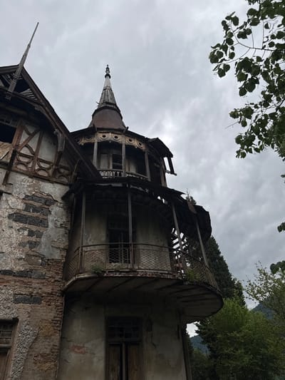 The image shows a dilapidated building in Akhtala, Armenia. The architecture suggests a historical structure, possibly a manor or a hotel, with elements of Victorian or Art Nouveau design. The building is in a severe state of disrepair, with crumbling plaster, exposed brickwork, and decaying wooden elements. A prominent feature is a multi-story turret with a conical roof, surrounded by balconies that are also in a state of deterioration, with rusted railings and missing sections. The windows are broken or boarded up, and the overall impression is one of abandonment and decay. The surrounding environment appears to be a natural landscape, with lush green trees and a cloudy sky. There are no people visible in the image.