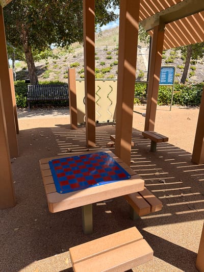 The image shows an outdoor recreational area in Irvine, United States of America, featuring a covered structure with wooden pillars and a slatted roof. Prominently displayed is a table with a blue and red checkerboard on its surface, suggesting a game area. Two benches are visible, one longer dark metal bench in the background and two shorter wooden benches closer to the foreground. The ground is covered with a fine gravel material. In the background, a grassy, sloped hill with scattered vegetation and trees is visible. A blue sign with text, likely playground rules, is mounted on a pole to the right of the structure. The scene is bathed in sunlight, casting distinct shadows from the roof onto the ground and the checkerboard table.
