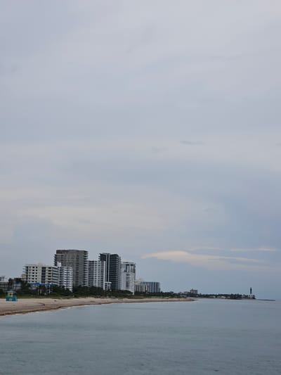 The image shows a view of the Pompano Beach coastline in Pompano Beach, Florida, USA. The scene is captured on an overcast day with a cloudy sky dominating the upper portion of the frame. In the mid-ground, a line of modern high-rise buildings stands against the horizon. These buildings are a mix of white and darker facades, with many featuring balconies. A sandy beach stretches along the foreground, meeting the calm, gray ocean. To the left, a small, brightly colored lifeguard station is visible on the beach. Further to the right, along the coastline, a lighthouse can be seen, indicating a more developed or historical area. The water is textured with gentle ripples. There are no discernible individuals engaged in specific actions within the frame, only the presence of the built environment and the natural elements of the sea and sky.