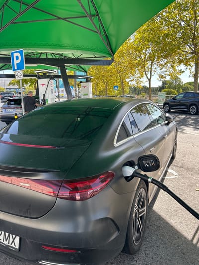 The image displays an IONITY electric vehicle charging station located at Continente Shopping in Leiria, Portugal, under clear skies. In the foreground, a dark grey electric sedan, likely a Mercedes-Benz EQE or EQS with a matte finish, is actively charging. A black charging cable is inserted into the vehicle's rear-right charging port, with a digital display visible on the charging handle. The car's partial Portuguese license plate, ending in "2MX", is visible. The vehicle is parked on asphalt with visible white parking bay lines, beneath a large green canopy supported by a dark metal structure.
In the mid-ground, a male individual wearing a dark t-shirt stands near an adjacent charging bay to the left, appearing to interact with a charging pedestal for a silver Audi sedan bearing the Portuguese license plate "8E 66 ZO". Multiple white IONITY charging pedestals are visible, featuring the brand's logo and green status indicators. A blue square parking sign with a white 'P' and an electric vehicle symbol is affixed to a pole under the canopy.
The background features numerous trees with yellow-green foliage, indicative of late summer or early autumn. A blue triangular pedestrian crossing sign is visible among the trees. Further to the right, a dark SUV is parked in the distance.