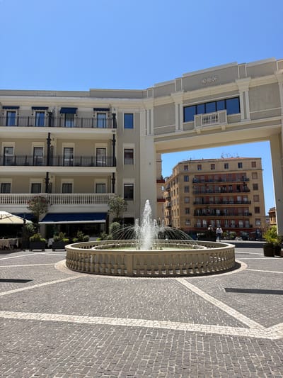 The image depicts a view of Piazza Yenne in Cagliari, Italy, on a clear, sunny day. In the foreground, a circular stone fountain with ornate balustrades is actively spraying water. The ground surrounding the fountain is paved with cobblestones laid in a geometric pattern with white stone accents. To the left of the fountain, part of an outdoor seating area for a restaurant is visible, with tables, chairs, and an umbrella.
Dominating the mid-ground is a multi-story building with a modern, neoclassical architectural style. It features balconies with black metal railings, blue awnings over some windows, and decorative cornices. This building forms an archway through which another building is visible.
In the background, beyond the archway, stands a taller, older apartment building with a pale yellow facade and numerous balconies adorned with red railings and air conditioning units. Two individuals are walking in front of this building. The sky above is a clear, bright blue. The scene captures a bustling urban square with a blend of contemporary and older architecture.
