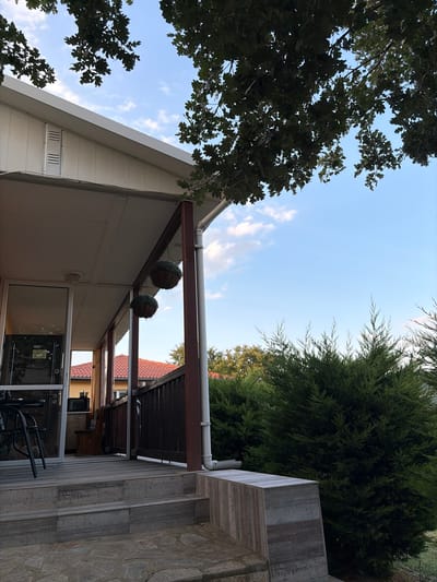 The image depicts a partial view of a house's exterior, likely a porch or veranda, in the city of Gniazdovo, Bulgaria. The foreground shows a set of grey tiled steps leading up to a wooden deck. To the left, a glass door and window reveal a glimpse of the interior, including a table, chairs, and what appears to be a small kitchen area with a microwave. Two hanging spherical planters with green foliage are suspended from the porch ceiling. A dark wooden railing lines the edge of the deck. In the background, a red-tiled roof of an adjacent building is visible, along with dense green trees and shrubs. The sky above is a clear blue with scattered white clouds. The overall scene suggests a residential property during daylight hours, with ample vegetation and architectural elements typical of a suburban or rural setting in Bulgaria. No people are visible in the image.