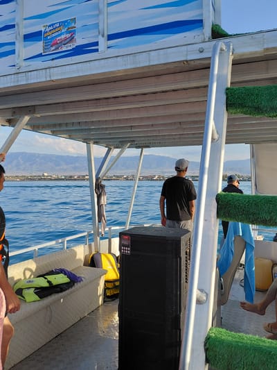 This media file depicts a group of people on a boat trip on a body of water, likely a lake, given the mountainous landscape visible in the background. The scene is set during the daytime, under a clear, bright sky, suggesting fair weather.
The primary subjects are several individuals, some of whom are partially visible or seen from behind. One person is standing near the front of the boat, facing away from the camera, wearing a black t-shirt and a grey baseball cap. Another person, also seen from the back, is wearing a blue tarp-like material draped over them. The boat itself appears to be a passenger vessel, with a covered section and metal railings. There are also various objects on board, including a large black speaker, yellow floatation devices, and a life vest.
A notable detail is a poster on the side of the boat with text in what appears to be a Cyrillic-based alphabet. The text includes "Аралға апарамыз" which translates to "We take you to Aral," and a phone number "77 633 44 77." This strongly suggests the boat is offering tours to the Aral Sea region. The overall atmosphere seems relaxed, consistent with a recreational boat trip. The water is calm and blue, reflecting the clear sky. The mountainous terrain in the distance provides a scenic backdrop.