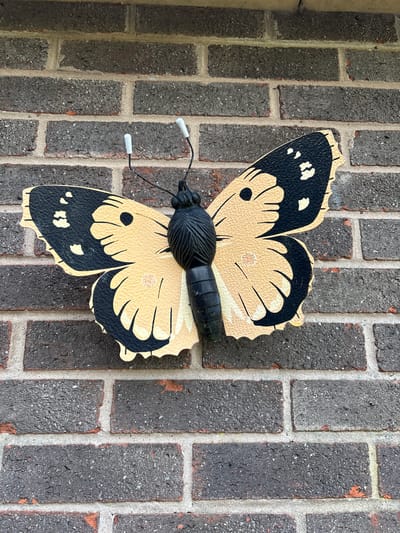 A close-up, eye-level shot reveals a decorative butterfly affixed to a brick wall. The butterfly is predominantly beige with black accents and white-tipped antennae. It is attached to the wall, which is made of dark grey bricks with lighter grout. The scene lacks any people, actions, or interactions. The image provides no information to confirm the location in Minsterley, United Kingdom.