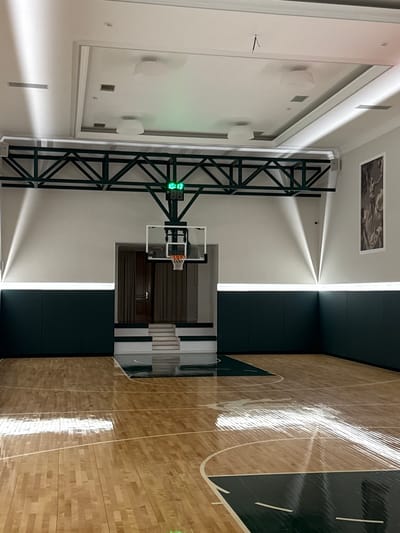 The image shows an indoor basketball court in Zhukovka, Russia. The court is well-lit with artificial lighting. A digital scoreboard displays "10:13". A basketball hoop with a transparent backboard is visible. The walls are painted white and dark green, and there are lights along the top of the dark green wainscoting. The wooden floor is polished and reflects the light. In the background, there are stairs leading up to a doorway with curtains. A framed artwork is hanging on the wall. The ceiling has recessed lighting and ventilation grilles. The overall environment appears to be a modern and well-maintained sports facility.