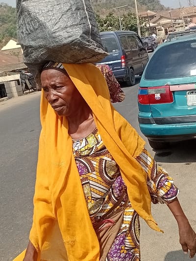 An elderly woman wearing a colorful patterned dress and a yellow headscarf carries a large gray sack on her head. She is walking on a paved road with vehicles parked beside her. The background shows a landscape of buildings and a distant hill under a bright, sunny sky.