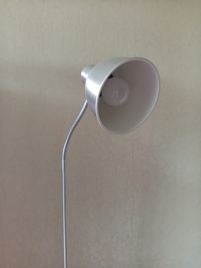 An indoor, close-up, medium shot shows a silver floor lamp against a textured, light beige wall. The lamp has a conical shade and a flexible neck. A white light bulb is visible inside the shade. The background wall appears to have a subtle, patterned texture. No people are present in the image. The image does not provide any information about the city of Vilyuysk, Russia.