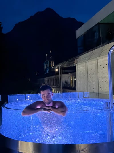 A man is submerged in a circular hot tub at night. The hot tub is illuminated with bright blue lights, creating a vibrant glow around the water. The man is leaning on his arms, which are crossed in front of him, and looking directly at the camera with a relaxed expression. The water appears bubbly and inviting.

In the background, a dark mountain range is silhouetted against a deep blue night sky. Below the mountains, some buildings are visible, one of which has a distinctive domed structure with illuminated windows. To the right of the frame, a modern building with a textured facade and glass elements is partially visible. A metal railing is also present, suggesting this is an outdoor amenity.

The scene takes place at night, as indicated by the dark sky and the artificial lighting. The weather appears clear, with no visible rain or clouds. The presence of a mountain range and European-style architecture suggests a mountainous region. The man's relaxed posture and the inviting atmosphere of the hot tub convey a sense of tranquility and enjoyment. There is no visible text in the image.