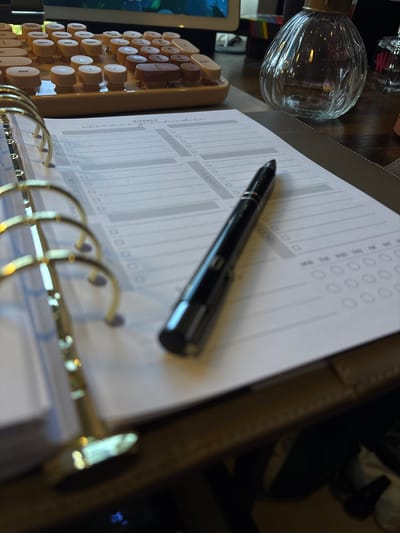 The image shows a close-up of a desk with various objects. A planner with gold rings lies open, displaying a page titled "WEEKLY cleaning schedule." The days of the week, Monday through Thursday, are printed on the page. A black pen rests on the page. To the left of the planner is a cream-colored mechanical keyboard with round keys, and behind that is a partially visible screen. A glass bottle-shaped object is also present, and a glimpse of a person or chair is visible in the upper right corner. The location is not identifiable as being in Ipswich, United Kingdom.