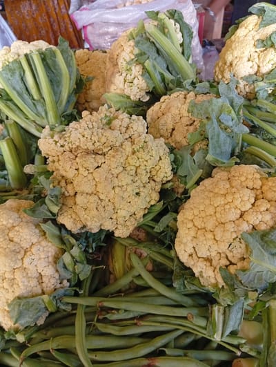 A close-up, high-angle shot displays a vibrant assortment of fresh vegetables, primarily cauliflower and green beans, at an outdoor market stall. The cauliflower heads, creamy white with hints of yellow, are piled generously, some still showing their leafy green crowns. Interspersed amongst the cauliflower are bundles of long, slender green beans, their smooth surfaces catching the light. The background is softly blurred, suggesting the bustling atmosphere of a market. In the upper portion of the image, a glimpse of a patterned fabric, possibly a sarong, and a white plastic bag with pink trim hints at the goods and packaging common in Indonesian markets. This scene likely depicts a section of the Meunyematangubi market in Indonesia, where fresh produce is a daily offering.