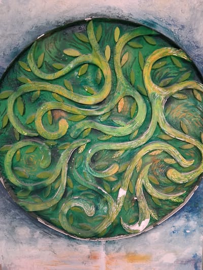 This is a close-up, overhead shot of a circular, bas-relief sculpture depicting an abstract, organic pattern of intertwined green vines and leaves. The sculpture is painted in varying shades of green and yellow, with some areas showing hints of brown and orange. The vines are thick and twisted, with a textured, almost bark-like appearance. Small, leaf-shaped forms are scattered throughout the design. The sculpture appears to be mounted or embedded in a textured background with washes of blue, white, and pink, suggesting it might be part of a larger wall art installation or a decorative element on a building. There are no people or animals visible in the image. The location in Constanța, Romania, is not discernible from the visual information provided in the image.