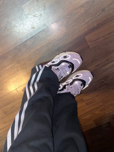 The image shows a person wearing gray athletic shoes with black and white detailing and black Adidas sweatpants with white stripes, standing on a hardwood floor. The location is not discernible from the image. No actions or interactions are visible.