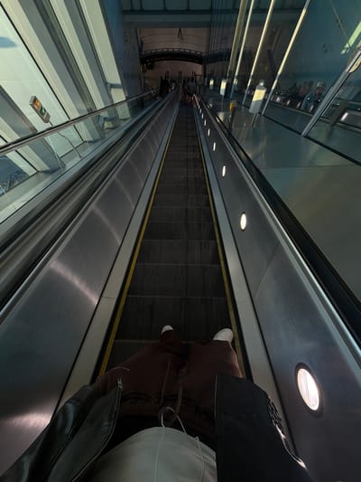 This is a first-person perspective image taken on an escalator at Gatwick Airport, Crawley, United Kingdom. The escalator is moving downwards, and the perspective is from someone standing on it, looking down at their own legs and the escalator steps. The escalator has metal sides and yellow safety lines. To the left, there's a glass balustrade and a modern architectural design with angled panels and lighting. On the right side of the escalator, there are recessed circular lights. In the distance, other people can be seen on the escalator and at the top. The overall environment suggests an airport terminal. The individual in the foreground is wearing a dark jacket, brown sweatpants, and white sneakers.