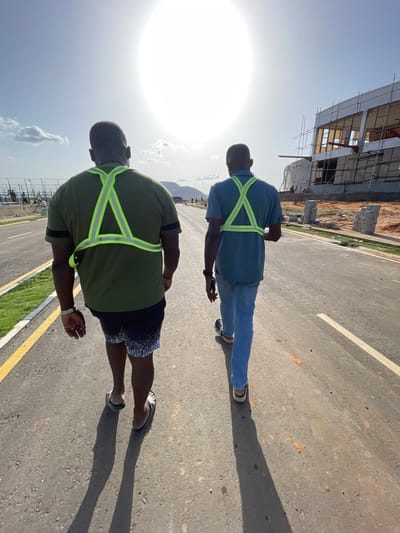Two men wearing safety vests are walking on a paved road. The road appears to be newly constructed, as there is adjacent construction visible in the background. The photograph was taken during the day in bright sunlight. The location is identified as Angwan Sayawa, Nigeria.