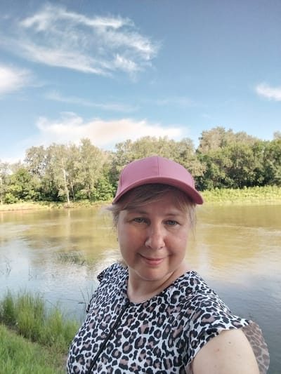 In this selfie, a woman with blonde hair and fair skin, wearing a pink baseball cap and a leopard-print blouse, is positioned in the foreground, looking towards the camera with a slight smile. She is situated beside a calm body of water, likely a river, with lush green trees and vegetation lining the opposite bank. The sky above is a clear blue with scattered wispy white clouds, indicating fair weather. The background features a dense treeline and a gentle ripple on the water's surface, suggesting a natural, serene environment. The location is implied to be the city of Chkalov, Russia, given the context.