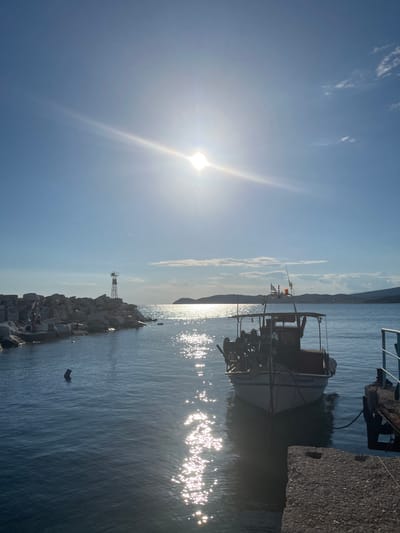 The image depicts a clear, sunny day at a harbor in Potos, Greece. The sun is high in the sky, creating a bright glare on the water. In the foreground, a concrete dock juts out into the sea. To the right of the dock, a small fishing boat is moored. The boat is white with blue trim and has a canopy over its deck. It is equipped with fishing gear, including reels and nets. In the middle ground, a rocky breakwater extends into the water, with a lighthouse-like structure at its end. The water is calm and blue, with sunlight reflecting off its surface. In the background, a distant coastline with hills is visible under a light blue sky with a few scattered clouds. The scene conveys a sense of peace and tranquility typical of a Greek coastal town.