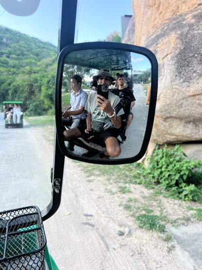 A mirror-image selfie is taken from the perspective of a passenger in a golf cart, capturing the scene ahead and to the side. The passenger, wearing a hat and a light-colored t-shirt, holds a phone to take the photo. Beside them, another passenger in a black t-shirt is visible. In the background, reflected in the mirror, a man in a light blue patterned shirt is seated, and other passengers are seen in the cart. Further ahead, another golf cart with passengers is driving on a dirt road. The environment features lush green hills on the left and a large rock formation on the right, suggesting a natural or park-like setting. The scene is likely set in Vĩnh Hải, Vietnam, given the context of the location.