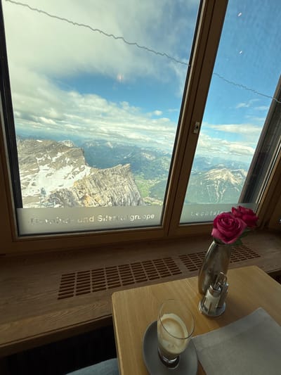 The photograph is taken from inside a building with a large window offering a panoramic view of a mountainous landscape. The window is framed by wooden beams. Through the window, a rugged mountain range with snow-capped peaks and green valleys is visible under a cloudy sky. In the foreground, on a wooden table, is a glass of coffee with crema, a salt and pepper shaker, and a vase with two pink roses. A grey napkin is placed next to the coffee. The text "Bernina- und Silvrettagruppe" and "Lechtaler" is visible etched on the lower part of the window, indicating a location in the Alps, likely near Ehrwald, Austria, given the reference to the Lechtal valley. The scene appears to be a serene moment in a cafe or restaurant with a stunning natural vista.