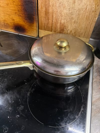 A close-up, eye-level shot captures a stainless steel pot with a gold knob, lid, and handle, sitting on a black glass stovetop. The pot's surface reflects the surrounding environment, with streaks and smudges indicating prior use. The stovetop itself is visibly dirty, with scattered white particles and circular heating element markings. Behind the pot, a wooden surface, likely a cutting board, shows signs of scorching and wear, with dark burn marks and wood grain details. The overall environment suggests a kitchen setting. The image does not contain information about the city of Crokhovo, Russia.