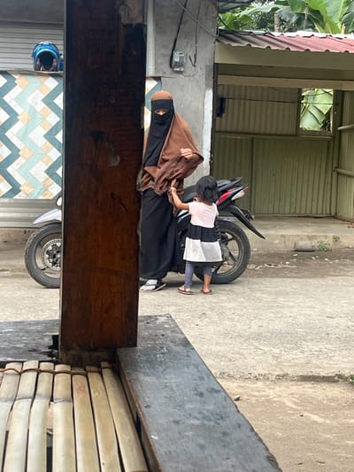 The image portrays an outdoor scene in Desa Batu Layar, Indonesia. A person wearing a brown head covering, black niqab, black long garment, and light-colored sandals stands on a concrete surface. Adjacent to this individual, a small child, dressed in a light pink, black, and white top, grey leggings, and brown flip-flops, holds onto the adult's garment with their left hand. A black and red motorcycle is parked directly behind the two individuals. To the left, a blue motorcycle helmet rests on a ledge against a building featuring a geometric tile pattern of light blue, white, and tan. A grey electrical meter is affixed to a concrete wall above the adult. To the right, a structure with a reddish-brown corrugated metal roof and light green corrugated metal walls is visible, with an open window or aperture. Green foliage is present in the upper right background. The foreground is partially obscured by a dark wooden pillar and a section of a bamboo-slatted bench.