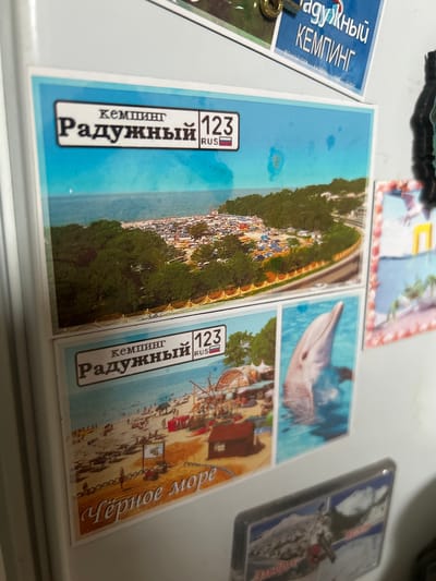 The image shows a collection of magnets on a refrigerator. The top magnet depicts an aerial view of a crowded beach or campsite with many tents and vehicles. Above the scene, text reads "кемпинг Радужный 123 RUS". A second magnet below shows a beach scene with people, umbrellas, and a Ferris wheel. The text on this magnet reads "кемпинг Радужный 123 RUS" and below that "Черное море". A third magnet shows a dolphin jumping out of the water. The fourth magnet at the bottom shows a mountain landscape with snow and a ski resort. The city of Primalkinskoye, Russia is not visible in the image.
