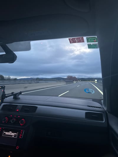 The image shows the view from the driver's seat of a car driving on a highway. The car is traveling in the left lane, and the road ahead appears clear with a few cars in the distance. The sky is overcast and gray. The highway is lined with trees and vegetation on both sides and there is a gray concrete barrier separating the road from the vegetation. The car's dashboard and the driver's side window are visible, including the steering wheel, the radio, and the gear stick. There is a smartphone attached to a holder on the dashboard. There is a sticker on the windshield with an unknown logo. There are no people visible in the image. The image was taken in La Losa, Spain, near the coordinates 40.842 latitude and -4.146 longitude.